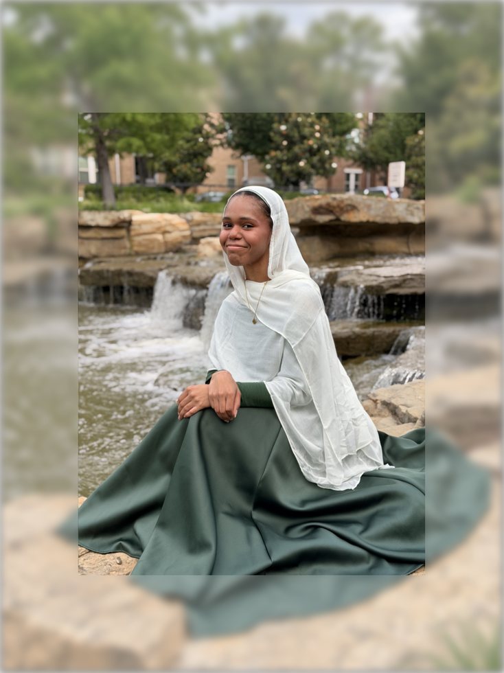 Faatimah sitting by a waterfall in a white scarf and green skirt