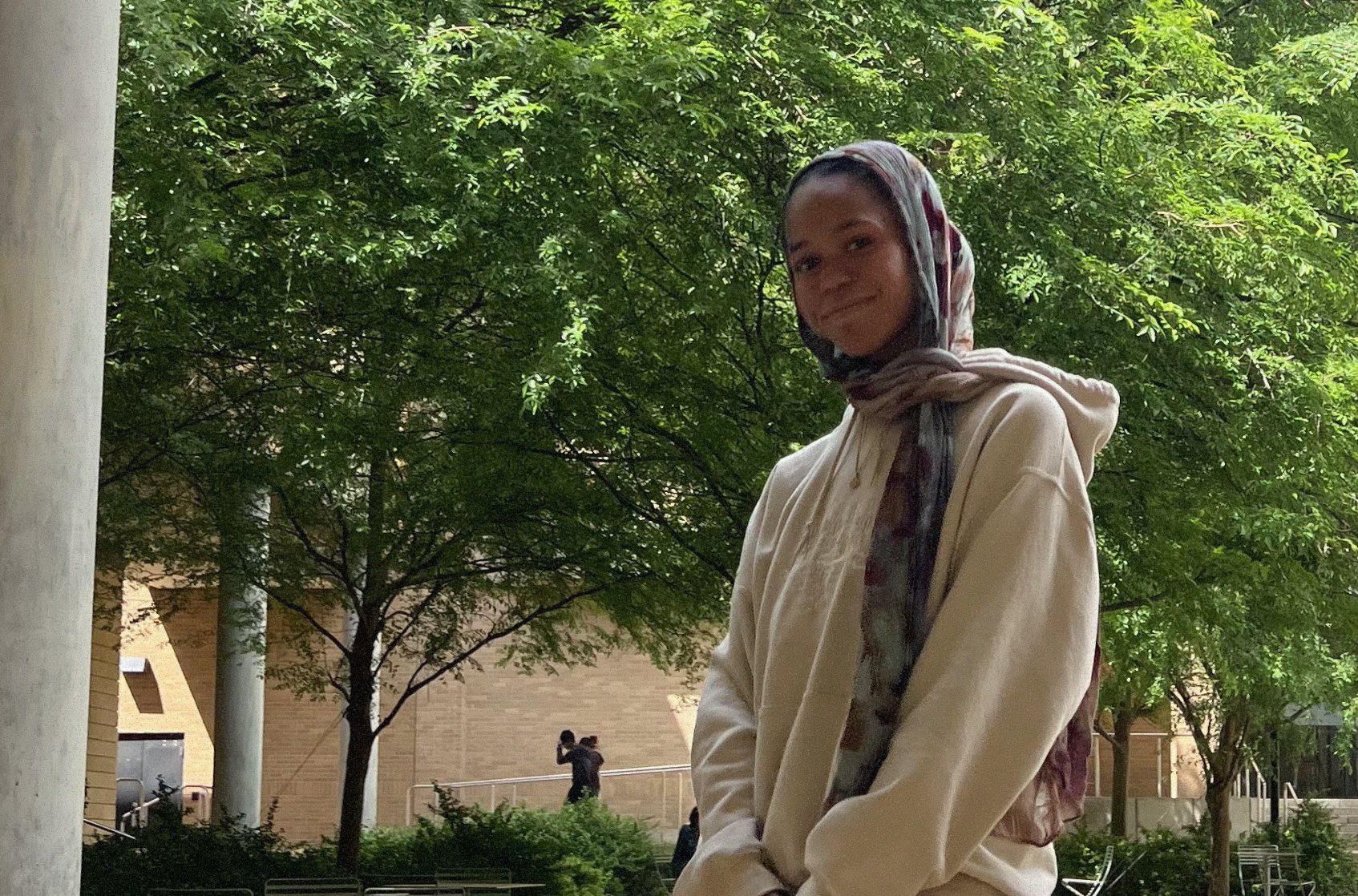 Faatimah standing under green trees in a cream hoodie and a patterned headscarf.